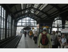 Eine überdachte Bahnhofsplattform mit Gleisen auf der linken Seite und einer Glasfassade, die viel Licht hereinlässt. Menschen stehen oder gehen entlang der Plattform, und ein Schild mit Ausgangsnummern ist sichtbar.