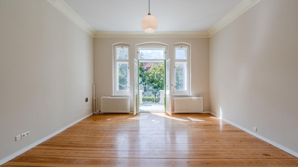 Leerer, heller Altbau-Wohnraum mit breiten Holzdielen, hohen Decken mit Stuck und zentraler Hängelampe. Am Ende des Raums befinden sich hohe Fenster und eine geöffnete Balkontür, flankiert von Heizkörpern, mit Blick ins Grüne.