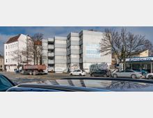 Straßenszene mit mehreren Gebäuden, darunter ein verputztes, weißes Wohnhaus mit sichtbaren Fassadenschäden und Graffiti sowie ein danebenliegendes Autosalon-Gebäude. Geparkte Autos, ein LKW und vereinzelte Fußgänger befinden sich auf dem Bürgersteig, während kahle Bäume die Umgebung säumen.