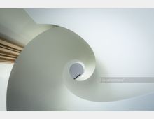 Ansicht einer weißen Wendeltreppe in moderner, minimalistischem Design aus einer ungewöhnlichen Perspektive, die den spiralartigen Verlauf und das zentrale Geländer betont. Am Rand ist ein Abschnitt einer Holzverkleidung zu sehen.