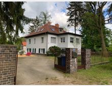 Zweistöckiges Wohnhaus mit weißer Fassade und rotem Satteldach, umgeben von einem Garten mit Bäumen. Im Vordergrund ist eine offene Einfahrt mit einem Eingangstor und Backsteinpfeilern zu sehen.