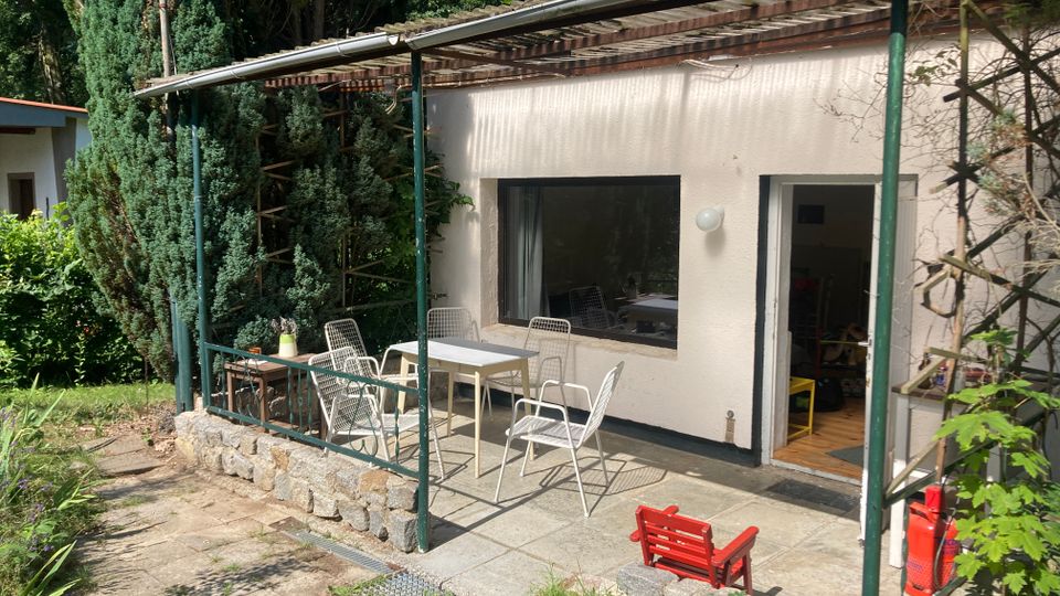 Überdachte Terrasse an einem eingeschossigen Haus mit weißer Fassade, großem Fenster und offener Tür. Auf dem gepflasterten Bereich stehen mehrere Metallstühle und Tische, umgeben von niedriger Steinmauer und dichtem Baumbestand im Garten.