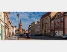 Straßenszene in einer Altstadt mit historischen Fassaden und einem markanten Backsteinturm einer Kirche im Hintergrund. Die Straße ist von geparkten Autos gesäumt und mit Kopfsteinpflaster belegt, während einige Laternen und Blumenkästen die Szene schmücken.