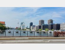Blick von einer Brücke auf die Spree in Berlin, mit modernen Bürogebäuden am Ufer und Baukränen im Hintergrund. Links ragt der Berliner Fernsehturm über die Skyline hinaus, während der Fluss im Vordergrund von einem schmiedeeisernen Brückengeländer gesäumt ist.