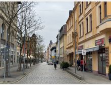 Eine gepflasterte Stadtstraße mit mehrstöckigen Gebäuden auf beiden Seiten, darunter Geschäfte und Restaurants, wie ein asiatisches Restaurant. Entlang der Straße stehen Bäume ohne Laub, und einige Passanten sind zu sehen.