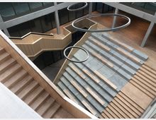 Offener Innenbereich eines modernen Gebäudes mit einer Treppenlandschaft aus Beton und Holzstufen, die als Sitzbereiche dienen. Über der Fläche hängen große, runde Leuchtelemente, und das Design kombiniert helle Holzelemente mit Glas- und Betonflächen.