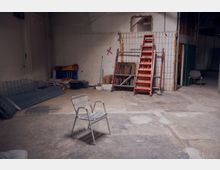 Ein leerer, unfertiger Raum mit sichtbaren Baustellenmaterialien, darunter eine rote Klappleiter, Metallgitter, Holzelemente und Eimer. Im Vordergrund steht ein einzelner Metallstuhl, die Wände sind teils gefliest und teilweise roh belassen.
