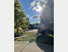 Ein Außenbereich mit Bäumen, einer zylinderförmigen, metallischen Struktur auf Stelzen und mehreren Tischen und Bänken im Hintergrund. Der Himmel ist sonnig mit einigen Wolken, und die Umgebung besteht aus Pflastersteinen, Gras und Beeten.