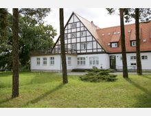 Fachwerkhaus mit weißer Fassade und dunkelbraunen Balken, umgeben von einer gepflegten Rasenfläche und hohen Bäumen; das Gebäude hat ein rotes Satteldach mit mehreren Dachfenstern und einen Balkon auf der linken Seite.