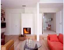 Modernes Wohnzimmer mit minimalistischer Einrichtung. Im Vordergrund befinden sich ein Glastisch und ein rotes Sofa, im Hintergrund ein Kamin mit Holzkorb daneben sowie eine offene Tür, die in einen weiteren Raum führt.