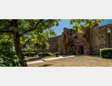 Mittelalterliche Stadtmauer aus Feldsteinen mit Backsteinelementen und einem Torbogen, umgeben von einem gepflegten Park mit Bäumen, Grasflächen und einem gepflasterten Weg. Im Hintergrund ist ein klarer blauer Himmel sichtbar.