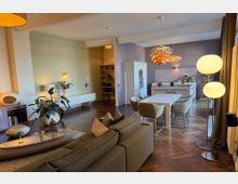 Open-plan modern living and dining room with a sofa and low coffee table leading to a long dining table and minimalist kitchen, all set on dark herringbone wood flooring. Pendant lights hang over the dining area, and large windows with curtains line one side of the room.