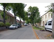 Ruhige, von Bäumen gesäumte Wohnstraße mit parkenden Autos und Häusern mit Satteldächern und heller Fassade auf beiden Straßenseiten. Im Vordergrund stehen ein weißer Kleinbus und ein Auto, während im Hintergrund die Straße in die Ferne führt.