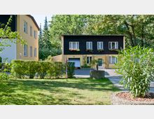 Wohnhaus in einer grünen Umgebung mit zwei Stockwerken, gelb gestrichenem Erdgeschoss und dunkler Holzfassade im oberen Bereich. Vor dem Gebäude befinden sich Beete mit Pflanzen, eine Hecke und ein gepflegter Rasen; im Hintergrund sind Bäume sichtbar.