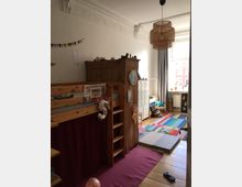 Ein helles Kinderzimmer mit Holz-Etagenbett, daran angehängter Basketballkorb und roter Vorhang. Im Hintergrund befinden sich bunte Spielmatten, eine weiße Kommode mit Schubladen und ein großes Fenster, das Tageslicht hereinlässt. Eine geflochtene Deckenlampe hängt zentral im Raum.