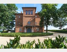 Backsteinbau mit symmetrischer Fassade und zentralem Haupteingang mit einem bogenförmigen, dunklen Tor, umgeben von gepflegten Hecken und Bäumen in einer parkähnlichen Umgebung. Im Vordergrund sind grüne Sträucher zu sehen, während der blaue Himmel die Szene im Hintergrund abrundet.