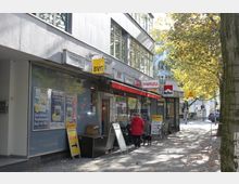 Straßenansicht eines kleinen Zeitungsladens mit Schaufenstern und mehreren Werbeschildern, darunter BVG, Lotto und Marlboro. Auf dem Gehweg stehen eine Person in roter Jacke mit Hund, ein Fahrrad und ein Baum im Vordergrund, während die straßenseitigen Gebäude von Herbstbäumen umgeben sind.