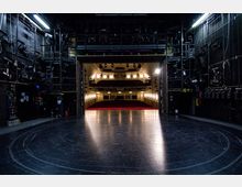 Blick von der Bühne eines Theatersaals in Richtung Zuschauerraum, mit sichtbaren Scheinwerfern und Bühnentechnik im Vordergrund. Der Zuschauerraum ist elegant gestaltet, mit roten Sitzreihen und Wandleuchten, die den Saal beleuchten.