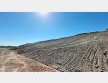 Eine aufgeschüttete, leicht geneigte Erdfläche mit sichtbaren Spuren von Baumaschinen und vereinzelter Vegetation am oberen Rand, unter wolkenlosem, blauem Himmel mit sichtbarer Sonneneinstrahlung. Im Hintergrund ist flaches Land erkennbar.
