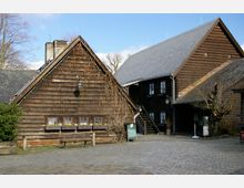 Holzgebäude in traditioneller Bauweise mit Satteldächern, Schindelverkleidung und kleinen Fenstern; vor dem Gebäude ein gepflasterter Hof sowie eine Sitzbank. Rankpflanzen bedecken teilweise die Fassade, und ein Hinweisschild steht an der Seite.