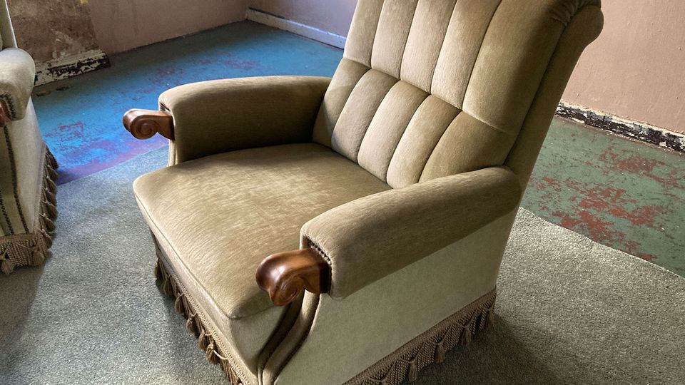 Innenraum eines älteren, teils abgenutzten Zimmers mit beigefarbenem Polstersessel mit Holzarmlehnen und Fransenbesatz. Der Boden ist mit grauem Teppich und sichtbaren blau-grünlichen, abgenutzten Flächen bedeckt; an den Wänden sind Gebrauchsspuren und eine Steckdose zu sehen.