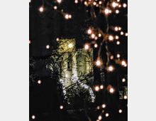 Ein beleuchtetes Gebäude bei Nacht, sichtbar durch Zweige und umgeben von kleinen, unscharfen Lichtern, die wahrscheinlich Teil von Weihnachtsdekorationen sind. Im Vordergrund sind glühende Lichtpunkte zu sehen, die den Blick auf das Gebäude teils verdecken.