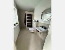 Modernes Badezimmer mit beigen Fliesen, einem wandhängenden WC und einem weißen Waschbecken, darüber ein runder Spiegel. Eine schwarze Handtuchheizung und kleine dekorative Elemente wie Pflanzen und Zahnbürstenhalter ergänzen die minimalistische Einrichtung.