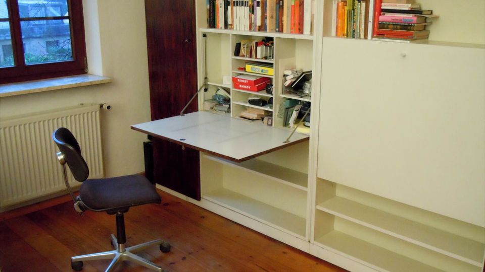 Arbeitszimmer mit großem weißen Einbauregal voller Bücher und einer ausklappbaren Schreibtischplatte. Daneben ein Fenster mit Heizkörper darunter sowie ein Holzfußboden und ein Bürostuhl vor dem Regal.