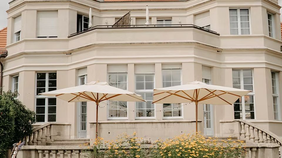 Große, helle Villa mit symmetrischer Fassade, hohen Fenstern und einer erhöhten Terrasse mit Balustrade und seitlichen Treppen. Im Vordergrund stehen auf einer Rasenfläche lange Tische mit Stühlen sowie zwei große Sonnenschirme vor einem Blumenbeet.