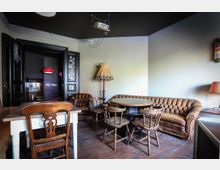 Gemütlicher Raum mit Vintage-Möbeln, darunter ein runder Holztisch mit Stühlen, ein gepolstertes Ledersofa und eine Stehlampe mit Rüschen. Im Hintergrund eine schwarze Türöffnung mit roter Hängelampe und Dekoelementen an den Wänden.