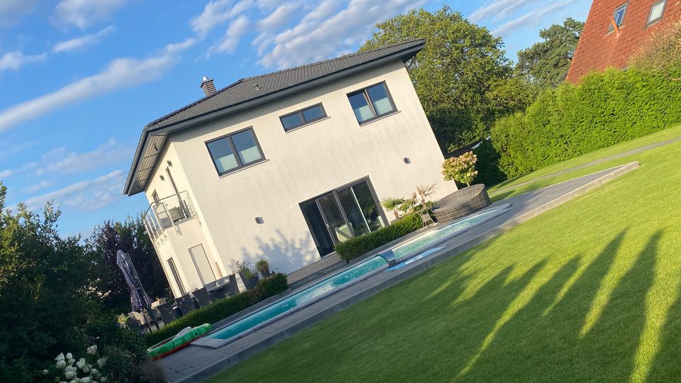 Modernes, zweistöckiges Wohnhaus mit heller Fassade und dunklem Satteldach, davor eine Terrasse mit langem, rechteckigem Pool. Im Vordergrund liegt ein gepflegter Rasen, umgeben von Hecken und Bäumen unter blauem Himmel.