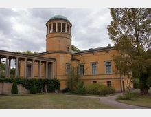 Gelbes, historisches Gebäude mit Säulengang und rundem Turm mit Kuppeldach. Die Umgebung besteht aus Rasenflächen, Bäumen und Büschen, unter einem bewölkten Himmel.