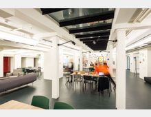 Moderner Gemeinschaftsraum mit einer offenen Gestaltung, weißen Säulen und dunklem Boden. Es gibt Sitzgruppen mit Sofas und Tischen, Arbeitsbereiche mit Stühlen und eine Glasdecke, die den Raum beleuchtet; bunte Poster an der Wand im Hintergrund.