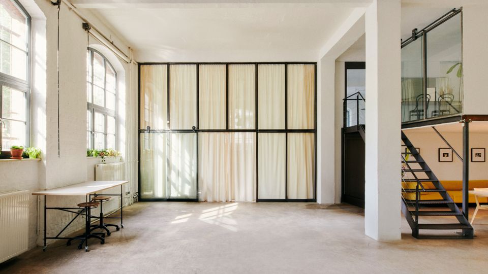 Heller, loftartiger Innenraum mit hohen Decken, großen Sprossenfenstern und einer schwarzen Glas-Trennwand mit Vorhängen. Rechts führt eine Metalltreppe zu einer Galerie mit Glasgeländer; links steht ein schlichter Arbeitstisch an der Fensterfront.