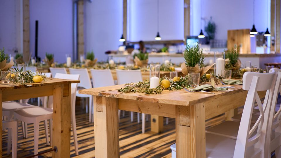 Ein modern eingerichteter Veranstaltungsraum mit Holztischen, weißen Stühlen und dekorativen Elementen wie Zitronen, Kerzen und grünem Laub auf den Tischen. Der Boden besteht aus gestreiftem Holz, während hängende Lampen und eine Bar im Hintergrund die Atmosphäre ergänzen.