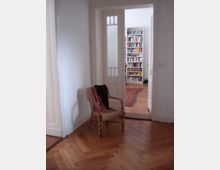 Ein Zimmer mit Parkettboden, einem geflochtenen Stuhl und einer offenen Tür, die in einen benachbarten Raum mit Bücherregalen und einem Teppich führt. Die Wände sind weiß, und die Tür besitzt Glaseinsätze.