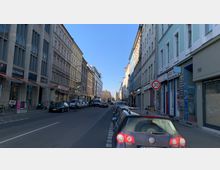 Städtische Straße mit mehrstöckigen Wohn- und Geschäftsgebäuden auf beiden Seiten, geparkte Autos am Straßenrand und Geschäfte im Erdgeschoss. Ein Verkehrsschild zeigt eine Tempo-30-Zone, während die Straße in der Ferne von einem größeren Gebäude abgeschlossen wird.