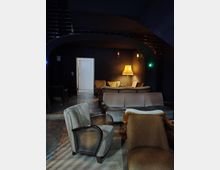 Gemütlicher Raum mit dunklen Wänden und einer Gewölbedecke, eingerichtet mit verschiedenen Polstermöbeln, darunter Sessel und Sofas. Eine Stehlampe mit gelbem Schirm beleuchtet den Hintergrund dezent, neben einer weißen Tür und mehreren Wandleuchten.