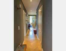 Ein schmaler, gut beleuchteter Hausflur mit grauen Wänden, Holzboden im Fischgrätmuster und weißen Türen. Am Ende des Flurs ist ein heller Raum mit einer Glastür und Fenstern sichtbar.