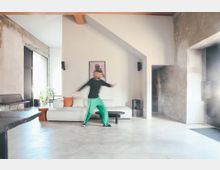 Wohnraum mit modernem und minimalistischem Design, geprägt durch Betonwände, eine helle Sofalandschaft mit bunten Kissen, große Fenster mit viel Tageslicht und offener Architektur. Im Vordergrund ist eine Person in Bewegung erkennbar.