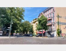 Straßenecke in einer Wohngegend mit Kopfsteinpflaster, umgeben von Bäumen und mehrstöckigen Gebäuden, darunter ein rosa verputztes Haus mit Balkonen. Im Vordergrund sind Straßenschilder, Laternen und geparkte Autos zu sehen.