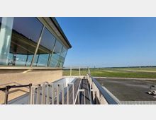 Außenansicht eines Gebäudes mit einer Glasfassade und Geländern, die zu einer Terrasse führen, im Vordergrund. Im Hintergrund erstreckt sich eine weitläufige Wiesenlandschaft mit vereinzeltem Baumbestand und Gebäuden am Horizont unter einem klaren blauen Himmel.