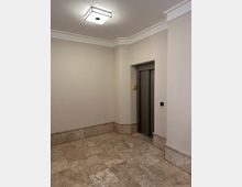 Kleines, helles Treppenhaus- oder Flurfoyer mit beigen Wänden, Steinfliesenboden und einer quadratischen Deckenleuchte. Rechts befindet sich eine eingelassene Aufzugstür mit Bedientaster an der Wand.