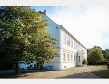 Weiße, zweistöckige Villa im klassischen Stil mit symmetrischen Fenstern und leicht hervorgehobenem Eingangsbereich. Im Vordergrund ein großer Baum, eine gepflasterte Fläche und eine Sitzbank, umgeben von einer grünen Parklandschaft.