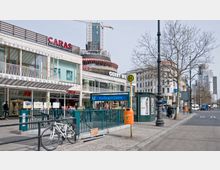 Straßenansicht am Kurfürstendamm in Berlin mit einem U-Bahneingang (Linien U1 und U9) im Vordergrund, Geschäftsgebäuden, darunter „Caras Gourmet Coffee“ und „H&M“, sowie einem Baustellenkran im Hintergrund. Neben dem Gehweg befinden sich Fahrradständer, eine Bushaltestelle und ein orangener Abfallbehälter.