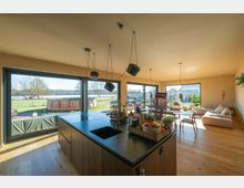 Moderne Wohnküche mit großem Küchenblock, Holzfußboden und hängenden Lampen. Große Fensterfronten bieten Blick auf eine Terrasse, eine grüne Wiese und einen runden Außenpool.