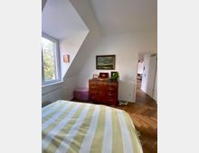 Schlafzimmer mit Holzfußboden und Dachschräge, einem Bett mit gestreifter gelb-weißer Bettwäsche, einer Kommode mit Dekorationsgegenständen und einem Gemälde darüber. Links ein Fenster mit Blick ins Freie, rechts eine geöffnete Tür zu einem weiteren Raum.