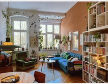 Wohnzimmer mit gemütlicher Einrichtung, darunter ein blaues Sofa, ein grüner Sessel und ein runder Teppich auf Holzboden. Die weißen Wände sind teilweise mit Pflanzen, Postern und Regalen voller Bücher dekoriert; große Fenster lassen viel Tageslicht herein.