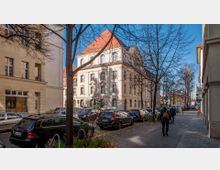 Straßenszene in einem städtischen Wohnviertel mit einem hellen, dreigeschossigen Gebäude im neoklassizistischen Stil und roten Dachziegeln im Hintergrund. Autos sind entlang der Straße geparkt, und vereinzelt gehen Passanten auf dem Gehweg, während kahle Bäume die Szene säumen.