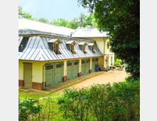 Gelbes Gebäude mit Satteldach und mehreren Dachgauben, darunter befinden sich grüne Garagentore in einer Reihe. Im Vordergrund sind Büsche und ein Schotterweg, daneben Fahrräder und ein kleiner Sitzbereich unter Bäumen.